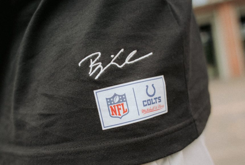 Eine Nahaufnahme einer Person, die ein schwarzes Hemd aus der Merchandise-Kollektion mit einer weißen Hose trägt. Das Hemd trägt Björn Werners Unterschrift über zwei Aufnähern: einem NFL-Logo-Aufnäher und einem Indianapolis Colts Mitchell & Ness-Aufnäher. Der Hintergrund ist unscharf. Diese Beschreibung wurde mit der FootballR KI automatisch generiert.