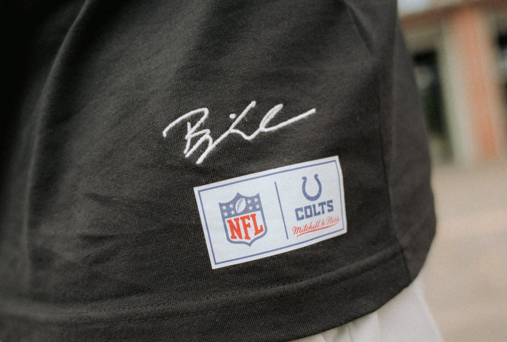 Eine Nahaufnahme einer Person, die ein schwarzes Hemd aus der Merchandise-Kollektion mit einer weißen Hose trägt. Das Hemd trägt Björn Werners Unterschrift über zwei Aufnähern: einem NFL-Logo-Aufnäher und einem Indianapolis Colts Mitchell & Ness-Aufnäher. Der Hintergrund ist unscharf. Diese Beschreibung wurde mit der FootballR KI automatisch generiert.