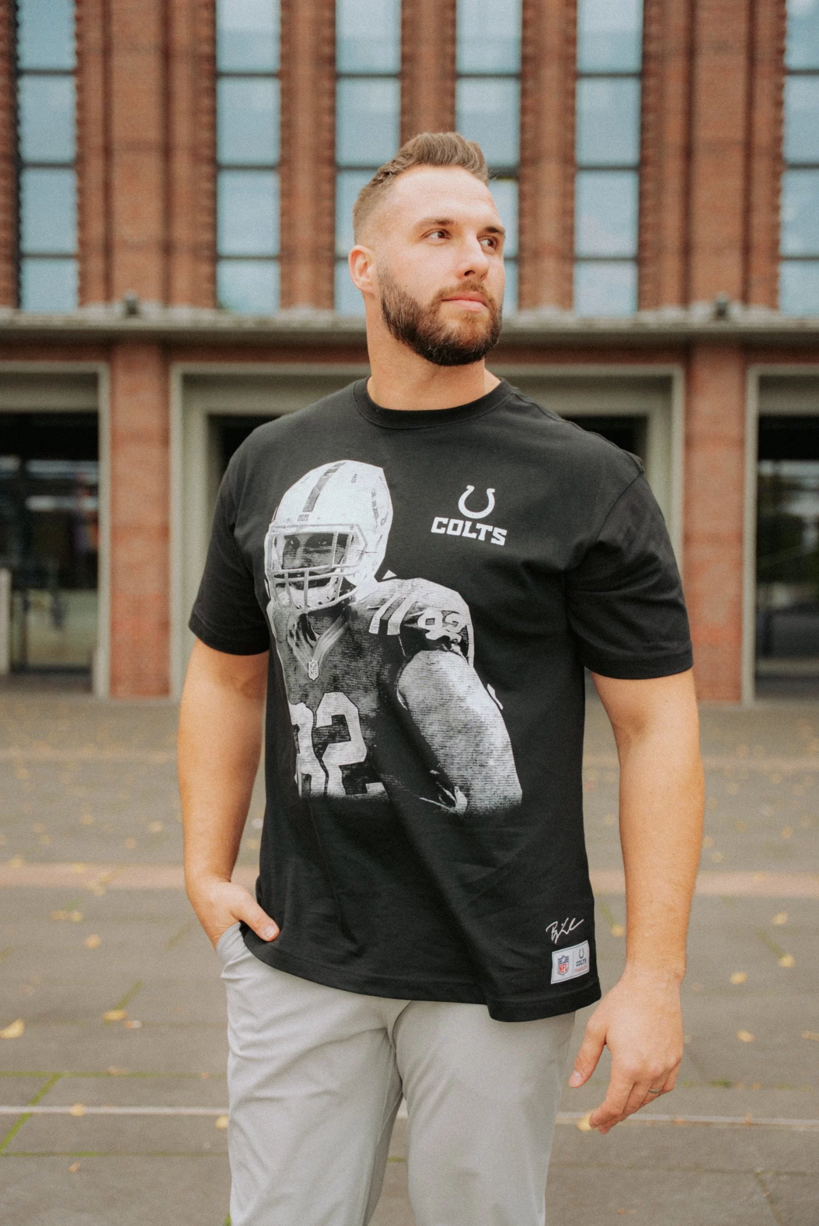 Ein bärtiger Mann steht im Freien vor einem Gebäude mit hohen Fenstern und trägt ein schwarzes T-Shirt der Indianapolis Colts Björn Werner Merchandise-Kollektion mit dem großen Graustufenbild eines Footballspielers. Eine Hand steckt in seiner Tasche, während er zur Seite blickt. Diese Beschreibung wurde mit der FootballR KI automatisch generiert.