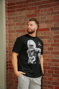 Ein bärtiger Mann mit kurzen Haaren, der ein schwarzes T-Shirt der Indianapolis Colts aus der Merchandise-Kollektion trägt, steht mit der rechten Hand in der Hosentasche vor einer roten Backsteinmauer und schaut leicht nach links. Diese Beschreibung wurde mit der FootballR KI automatisch generiert.