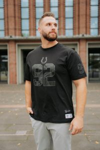 Ein bärtiger Mann steht im Freien vor einem Backsteingebäude. Er trägt ein schwarzes T-Shirt der Indianapolis Colts mit einer großen "92" und einer Hufeisengrafik - Teil der Merchandise-Kollektion von Björn Werner - und eine graue Hose. Er schaut mit einer Hand in der Tasche nach links. Diese Beschreibung wurde mit der FootballR KI automatisch generiert.