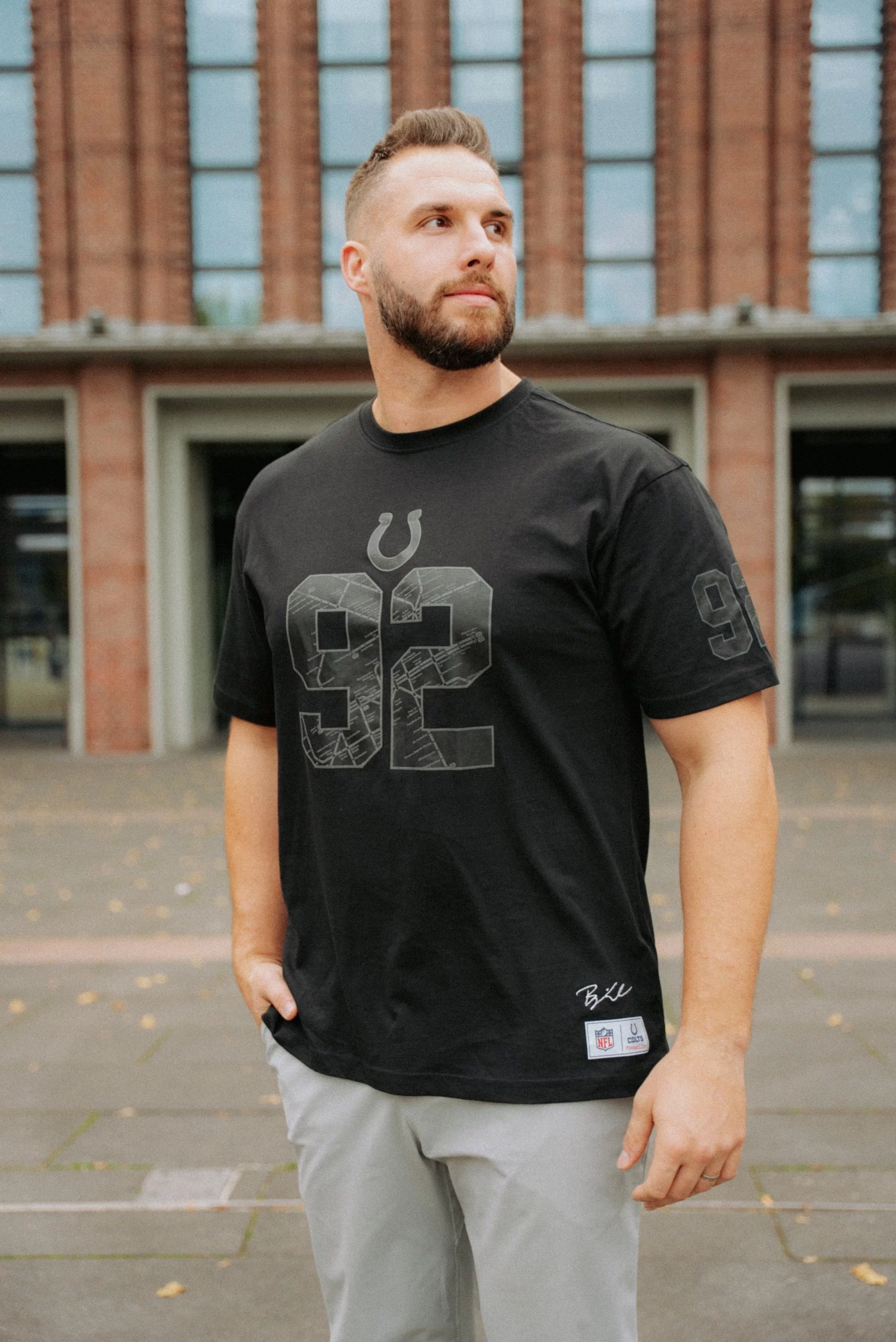 Ein bärtiger Mann steht im Freien vor einem Backsteingebäude. Er trägt ein schwarzes T-Shirt der Indianapolis Colts mit einer großen "92" und einer Hufeisengrafik - Teil der Merchandise-Kollektion von Björn Werner - und eine graue Hose. Er schaut mit einer Hand in der Tasche nach links. Diese Beschreibung wurde mit der FootballR KI automatisch generiert.