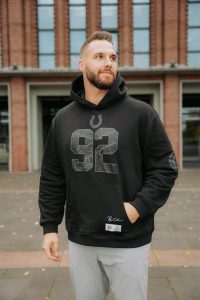 Ein bärtiger Mann steht im Freien vor einem Backsteingebäude und trägt einen schwarzen Indianapolis Colts-Kapuzenpullover aus der Björn Werner Merchandise-Kollektion. Seine linke Hand steckt in der Hosentasche, er schaut leicht zur Seite und lächelt sanft zwischen verstreuten Blättern. Diese Beschreibung wurde mit der FootballR KI automatisch generiert.