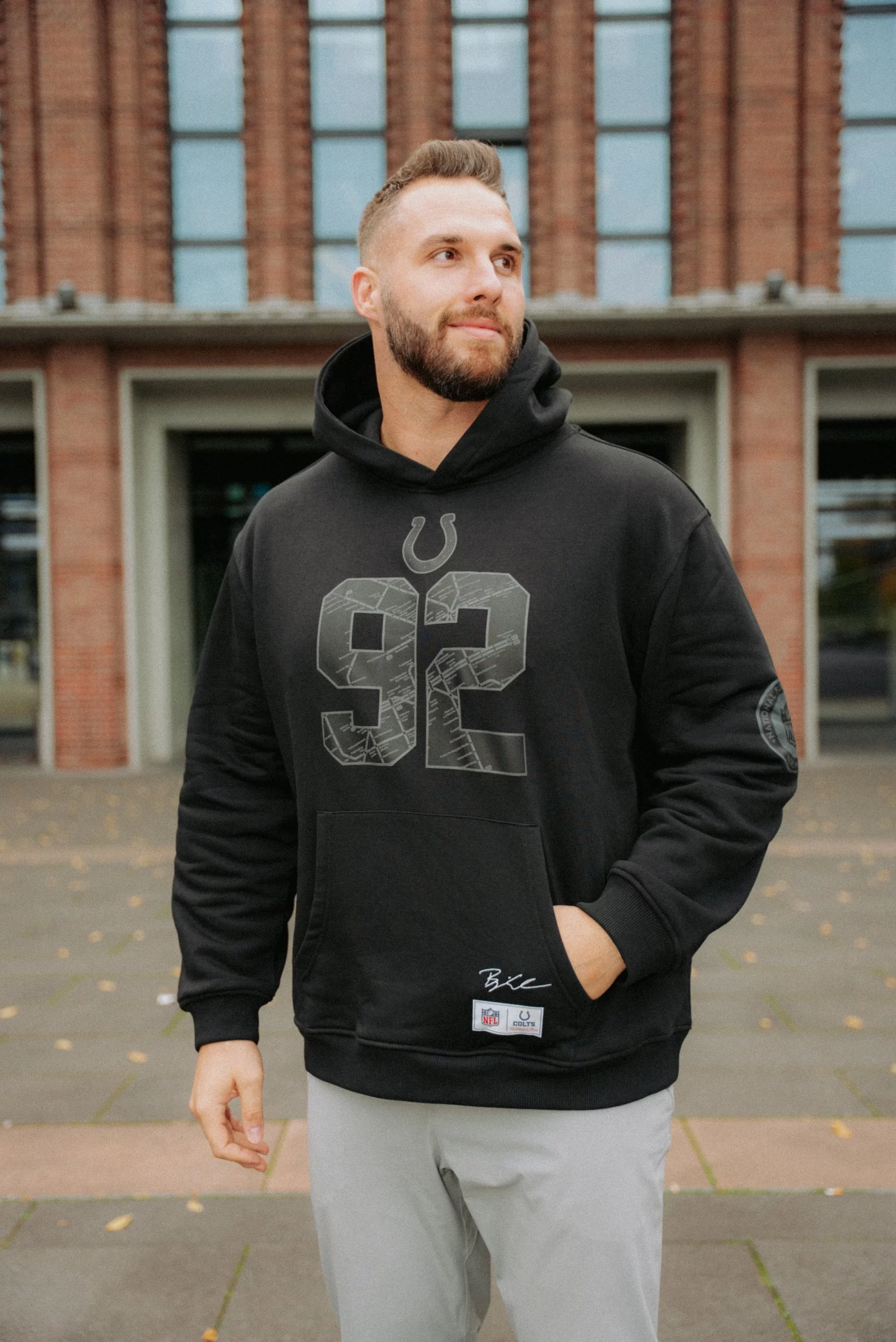 Ein bärtiger Mann steht im Freien vor einem Backsteingebäude und trägt einen schwarzen Indianapolis Colts-Kapuzenpullover aus der Björn Werner Merchandise-Kollektion. Seine linke Hand steckt in der Hosentasche, er schaut leicht zur Seite und lächelt sanft zwischen verstreuten Blättern. Diese Beschreibung wurde mit der FootballR KI automatisch generiert.