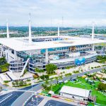 Bye Week 12 - Hard Rock Stadium - Luftaufnahme eines großen Stadions mit weißem Dach und hohen Stützmasten, mit Parkplätzen, Palmen und Grünflächen. Das Stadion brummt, während sich das topgesetzte Team auf sein Freilos in Woche 12 freut. In der Ferne ist die Skyline der Stadt bei teilweise bewölktem Himmel zu sehen. Diese Beschreibung wurde mit der FootballR KI automatisch generiert.