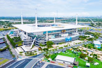 Bye Week 12 - Hard Rock Stadium - Luftaufnahme eines großen Stadions mit weißem Dach und hohen Stützmasten, mit Parkplätzen, Palmen und Grünflächen. Das Stadion brummt, während sich das topgesetzte Team auf sein Freilos in Woche 12 freut. In der Ferne ist die Skyline der Stadt bei teilweise bewölktem Himmel zu sehen. Diese Beschreibung wurde mit der FootballR KI automatisch generiert.