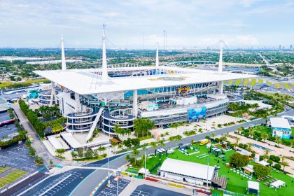 Bye Week 12 - Hard Rock Stadium - Luftaufnahme eines großen Stadions mit weißem Dach und hohen Stützmasten, mit Parkplätzen, Palmen und Grünflächen. Das Stadion brummt, während sich das topgesetzte Team auf sein Freilos in Woche 12 freut. In der Ferne ist die Skyline der Stadt bei teilweise bewölktem Himmel zu sehen. Diese Beschreibung wurde mit der FootballR KI automatisch generiert.