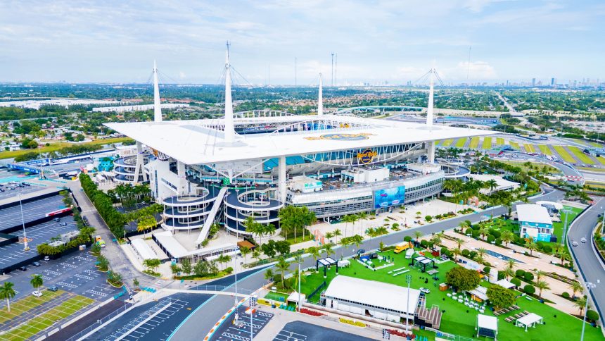Bye Week 12 - Hard Rock Stadium - Luftaufnahme eines großen Stadions mit weißem Dach und hohen Stützmasten, mit Parkplätzen, Palmen und Grünflächen. Das Stadion brummt, während sich das topgesetzte Team auf sein Freilos in Woche 12 freut. In der Ferne ist die Skyline der Stadt bei teilweise bewölktem Himmel zu sehen. Diese Beschreibung wurde mit der FootballR KI automatisch generiert.
