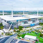 Bye Week 12 - Hard Rock Stadium - Luftaufnahme eines großen Stadions mit weißem Dach und hohen Stützmasten, mit Parkplätzen, Palmen und Grünflächen. Das Stadion brummt, während sich das topgesetzte Team auf sein Freilos in Woche 12 freut. In der Ferne ist die Skyline der Stadt bei teilweise bewölktem Himmel zu sehen. Diese Beschreibung wurde mit der FootballR KI automatisch generiert.