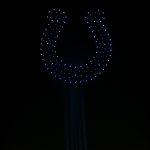 Der Nachthimmel über einem Berliner Stadion zeigt eine große Hufeisenform, die von vielen beleuchteten Drohnen in einer spektakulären Drohnenshow gebildet wird. Helle Lichtstrahlen erheben sich aus dem schwach beleuchteten Stadion und schaffen eine dramatische, hochtechnologische Atmosphäre. Diese Beschreibung wurde mit der FootballR KI automatisch generiert.