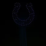 Der Nachthimmel über einem Berliner Stadion zeigt eine große Hufeisenform, die von vielen beleuchteten Drohnen in einer spektakulären Drohnenshow gebildet wird. Helle Lichtstrahlen erheben sich aus dem schwach beleuchteten Stadion und schaffen eine dramatische, hochtechnologische Atmosphäre. Diese Beschreibung wurde mit der FootballR KI automatisch generiert.