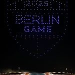 Nachts über einem Stadion in Berlin bildet eine atemberaubende Drohnenshow ein leuchtendes Schild mit der Aufschrift "2024 BERLIN GAME" in blauen und weißen Lichtern. Das beleuchtete Stadion ist voll mit Zuschauern, die Fahnen säumen das Dach. Diese Beschreibung wurde mit der FootballR KI automatisch generiert.