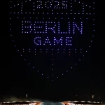Nachts über einem Stadion in Berlin bildet eine atemberaubende Drohnenshow ein leuchtendes Schild mit der Aufschrift "2024 BERLIN GAME" in blauen und weißen Lichtern. Das beleuchtete Stadion ist voll mit Zuschauern, die Fahnen säumen das Dach. Diese Beschreibung wurde mit der FootballR KI automatisch generiert.