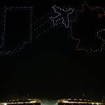 Eine nächtliche Drohnenshow erhellt den Himmel über einem Stadion, wobei Drohnen die Form des Bundesstaates Indiana in Blau, ein Colts-Flugzeug in Weiß und ein Rennwagenhelm in Rot und Orange nachbilden - eine Reminiszenz an die schillernden Flugvorführungen in Berlin. Diese Beschreibung wurde mit der FootballR KI automatisch generiert.