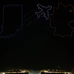 Eine nächtliche Drohnenshow erhellt den Himmel über einem Stadion, wobei Drohnen die Form des Bundesstaates Indiana in Blau, ein Colts-Flugzeug in Weiß und ein Rennwagenhelm in Rot und Orange nachbilden - eine Reminiszenz an die schillernden Flugvorführungen in Berlin. Diese Beschreibung wurde mit der FootballR KI automatisch generiert.
