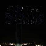 Nachts erhellt eine Drohnenshow den Berliner Himmel, während Drohnen in leuchtendem Blau "FOR THE SHOE" über einem glühenden Stadion buchstabieren. Vier vertikale Balken ragen nach oben und verstärken die dramatische Hommage an die Colts in der umgebenden Dunkelheit. Diese Beschreibung wurde mit der FootballR KI automatisch generiert.