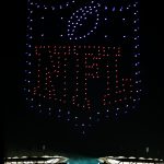 Ein nächtlicher Himmel über einem hell erleuchteten Stadion in Berlin bietet eine atemberaubende Drohnenshow, bei der Hunderte von beleuchteten Drohnen das NFL-Schildlogo über den Tribünen und Zuschauern formen; "NFL" leuchtet in roten Lichtern und hebt sich blau von der Dunkelheit ab. Diese Beschreibung wurde mit der FootballR KI automatisch generiert.