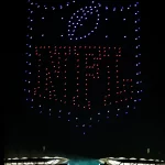 Ein nächtlicher Himmel über einem hell erleuchteten Stadion in Berlin bietet eine atemberaubende Drohnenshow, bei der Hunderte von beleuchteten Drohnen das NFL-Schildlogo über den Tribünen und Zuschauern formen; "NFL" leuchtet in roten Lichtern und hebt sich blau von der Dunkelheit ab. Diese Beschreibung wurde mit der FootballR KI automatisch generiert.
