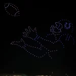 Ein nächtlicher Himmel über einem Berliner Stadion zeigt Hunderte von bunten Drohnen, die den Umriss eines Colts-Footballspielers bilden, der einen Football fangen will, beides in blauem und weißem Licht. Das beleuchtete Stadion und der dunkle Himmel bilden eine eindrucksvolle Drohnenshow-Kulisse. Diese Beschreibung wurde mit der FootballR KI automatisch generiert.