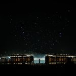 Ein hell erleuchtetes Stadion im nächtlichen Berlin ist von Dunkelheit umgeben. Links scheint der Vollmond, während über dem Stadion eine spektakuläre Drohnenshow bunte Muster bildet. Silhouettierte Menschen und Colts-Fans stehen im Vordergrund und lassen sich von der Szene faszinieren. Diese Beschreibung wurde mit der FootballR KI automatisch generiert.