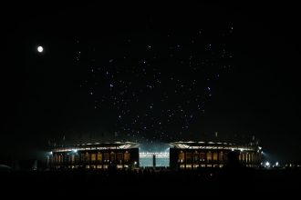 Ein hell erleuchtetes Stadion im nächtlichen Berlin ist von Dunkelheit umgeben. Links scheint der Vollmond, während über dem Stadion eine spektakuläre Drohnenshow bunte Muster bildet. Silhouettierte Menschen und Colts-Fans stehen im Vordergrund und lassen sich von der Szene faszinieren. Diese Beschreibung wurde mit der FootballR KI automatisch generiert.