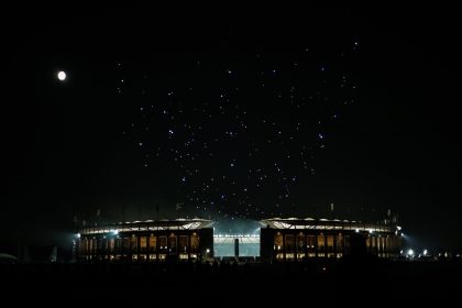 Ein hell erleuchtetes Stadion im nächtlichen Berlin ist von Dunkelheit umgeben. Links scheint der Vollmond, während über dem Stadion eine spektakuläre Drohnenshow bunte Muster bildet. Silhouettierte Menschen und Colts-Fans stehen im Vordergrund und lassen sich von der Szene faszinieren. Diese Beschreibung wurde mit der FootballR KI automatisch generiert.
