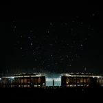 Ein hell erleuchtetes Stadion im nächtlichen Berlin ist von Dunkelheit umgeben. Links scheint der Vollmond, während über dem Stadion eine spektakuläre Drohnenshow bunte Muster bildet. Silhouettierte Menschen und Colts-Fans stehen im Vordergrund und lassen sich von der Szene faszinieren. Diese Beschreibung wurde mit der FootballR KI automatisch generiert.