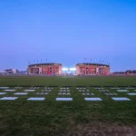 Ein weites, grasbewachsenes Feld führt zu einem großen, mit Fahnen geschmückten Stadion. Der Himmel ist klar und geht in der Nähe des Horizonts von Hellblau in Lila über. Auf dem Spielfeld sind riesige Schachfiguren aufgestellt - vielleicht für eine Veranstaltung zum Thema Colts oder eine Berliner Drohnenshow. Diese Beschreibung wurde mit der FootballR KI automatisch generiert.