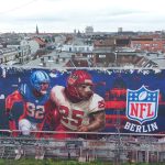Ein großes Wandgemälde auf einem städtischen Gebäude zeigt zwei American-Football-Spieler in Aktion neben einem leuchtenden NFL-Berlin Logo, das die Football-Kultur feiert. Das rot-blaue Stadtbild zeigt die Dächer, Antennen und Kräne Berlins hinter dem Wandbild. Diese Beschreibung wurde mit der FootballR KI automatisch generiert.