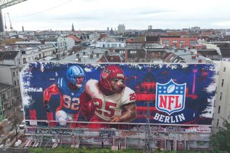 Ein großes Wandgemälde auf einem städtischen Gebäude zeigt zwei American-Football-Spieler in Aktion neben einem leuchtenden NFL-Berlin Logo, das die Football-Kultur feiert. Das rot-blaue Stadtbild zeigt die Dächer, Antennen und Kräne Berlins hinter dem Wandbild. Diese Beschreibung wurde mit der FootballR KI automatisch generiert.