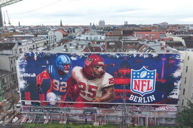 Ein großes Wandgemälde auf einem städtischen Gebäude zeigt zwei American-Football-Spieler in Aktion neben einem leuchtenden NFL-Berlin Logo, das die Football-Kultur feiert. Das rot-blaue Stadtbild zeigt die Dächer, Antennen und Kräne Berlins hinter dem Wandbild. Diese Beschreibung wurde mit der FootballR KI automatisch generiert.