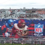 Ein großes Wandgemälde auf einem städtischen Gebäude zeigt zwei American-Football-Spieler in Aktion neben einem leuchtenden NFL-Berlin Logo, das die Football-Kultur feiert. Das rot-blaue Stadtbild zeigt die Dächer, Antennen und Kräne Berlins hinter dem Wandbild. Diese Beschreibung wurde mit der FootballR KI automatisch generiert.