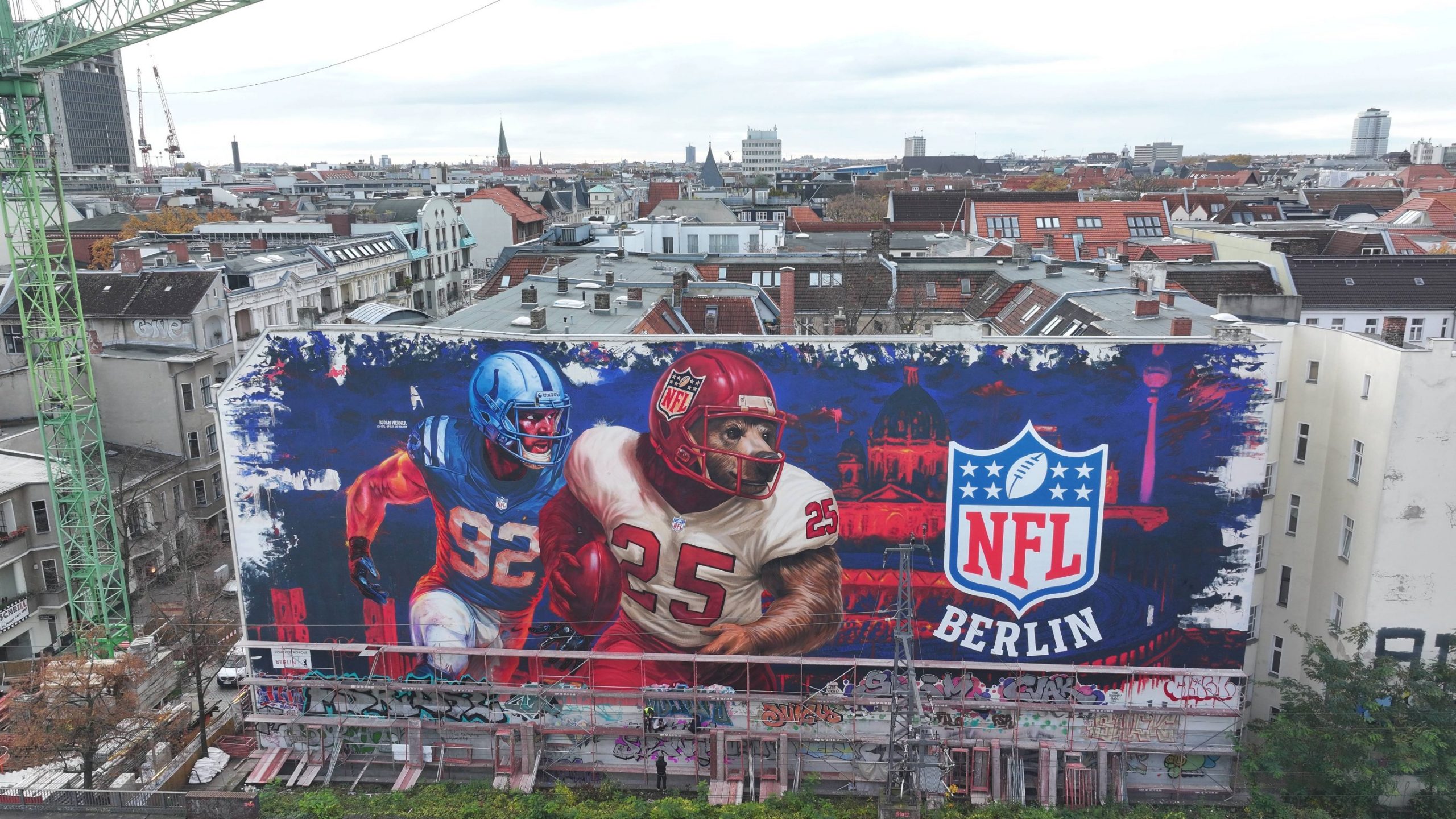 Ein großes Wandgemälde auf einem städtischen Gebäude zeigt zwei American-Football-Spieler in Aktion neben einem leuchtenden NFL-Berlin Logo, das die Football-Kultur feiert. Das rot-blaue Stadtbild zeigt die Dächer, Antennen und Kräne Berlins hinter dem Wandbild. Diese Beschreibung wurde mit der FootballR KI automatisch generiert.
