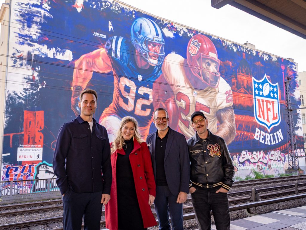 Vier Menschen stehen lächelnd vor einem lebendigen Wandbild, das Football-Kultur feiert und zwei American-Football-Spieler sowie das Logo der NFL Berlin zeigt. Graffiti bedecken die Wand und im Vordergrund verlaufen Bahngleise, die die urbane Energie Berlins während der NFL-Woche einfangen. Diese Beschreibung wurde mit der FootballR KI automatisch generiert.