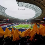 Woche 10 Recap - Ein vollbesetztes Berliner Stadion mit einem geschwungenen Dach und einem Fußballfeld in der Mitte strotzt vor Energie. Die Fans schwenken gelbe Fahnen im Vordergrund, während andere rote, weiße und schwarze Karten in der Hand halten und so ein farbenfrohes Bild unter einem bewölkten Himmel schaffen. Diese Beschreibung wurde mit der FootballR KI automatisch generiert.