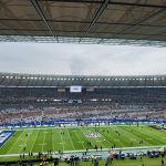 Ein großes Football-Stadion mit einem teilweise gefüllten Zuschauerraum und einem grünen NFL-Feld. Spieler und Betreuer stehen an der Seitenlinie, und eine Anzeigetafel zeigt "BAL" an. Der Himmel ist bedeckt, und die Tribünen sind rot, weiß und grau mit einigen verstreuten Fans. Diese Beschreibung wurde mit der FootballR KI automatisch generiert.