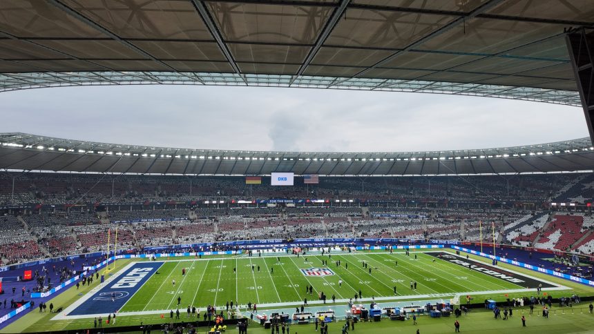 Ein großes Football-Stadion mit einem teilweise gefüllten Zuschauerraum und einem grünen NFL-Feld. Spieler und Betreuer stehen an der Seitenlinie, und eine Anzeigetafel zeigt "BAL" an. Der Himmel ist bedeckt, und die Tribünen sind rot, weiß und grau mit einigen verstreuten Fans. Diese Beschreibung wurde mit der FootballR KI automatisch generiert.