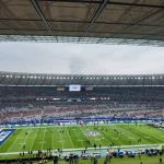 Ein großes Football-Stadion mit einem teilweise gefüllten Zuschauerraum und einem grünen NFL-Feld. Spieler und Betreuer stehen an der Seitenlinie, und eine Anzeigetafel zeigt "BAL" an. Der Himmel ist bedeckt, und die Tribünen sind rot, weiß und grau mit einigen verstreuten Fans. Diese Beschreibung wurde mit der FootballR KI automatisch generiert.