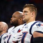 Drei American-Football-Spieler stehen Seite an Seite in den Trikots der New England Patriots und blicken mit ernster Miene nach vorne. Rob Gronkowski steht im Vordergrund, ein echter Patriot, der während der Nationalhymne seine rechte Hand über sein Herz hält. Diese Beschreibung wurde mit der FootballR KI automatisch generiert.