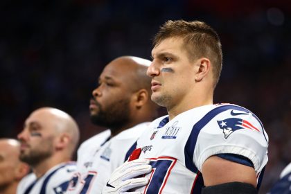 Drei American-Football-Spieler stehen Seite an Seite in den Trikots der New England Patriots und blicken mit ernster Miene nach vorne. Rob Gronkowski steht im Vordergrund, ein echter Patriot, der während der Nationalhymne seine rechte Hand über sein Herz hält. Diese Beschreibung wurde mit der FootballR KI automatisch generiert.