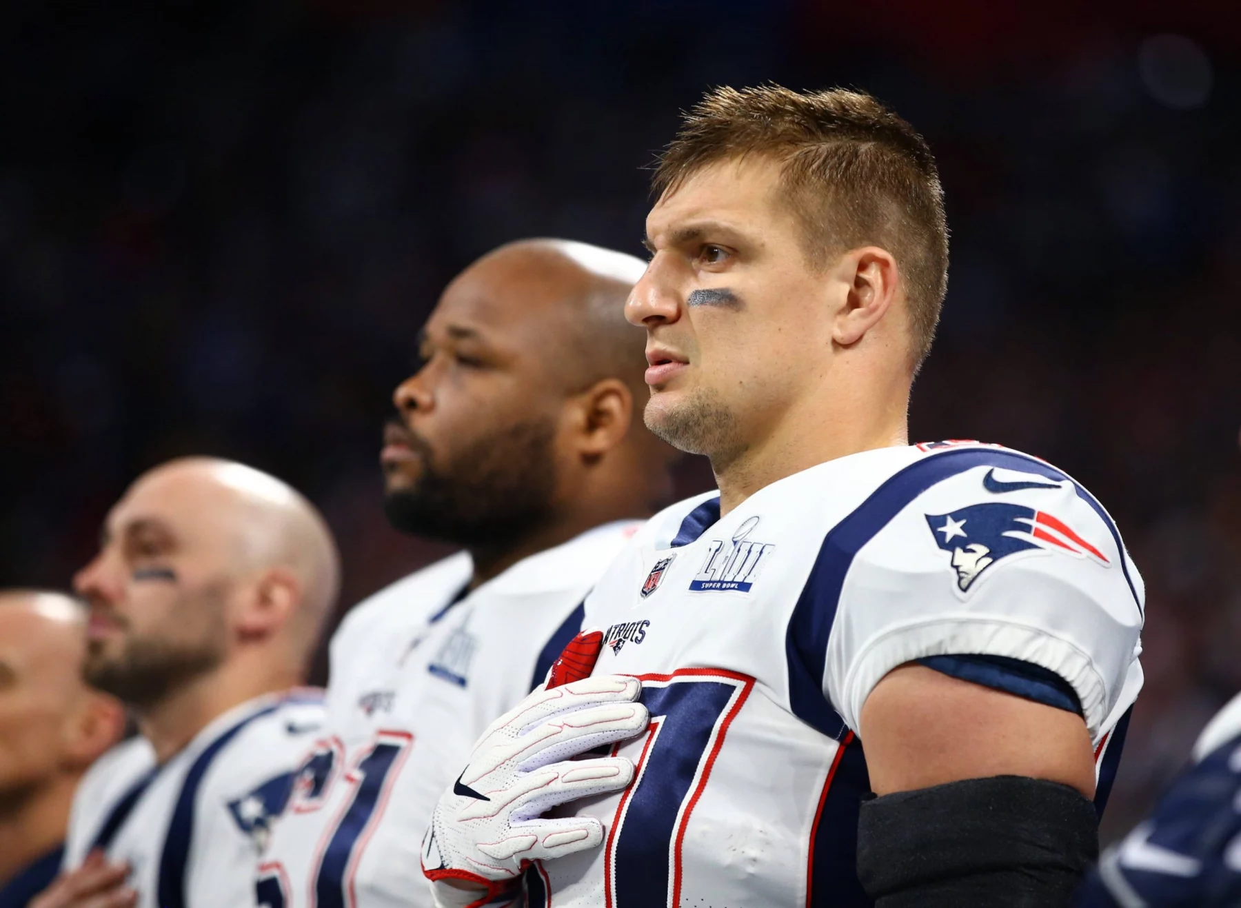 Drei American-Football-Spieler stehen Seite an Seite in den Trikots der New England Patriots und blicken mit ernster Miene nach vorne. Rob Gronkowski steht im Vordergrund, ein echter Patriot, der während der Nationalhymne seine rechte Hand über sein Herz hält. Diese Beschreibung wurde mit der FootballR KI automatisch generiert.