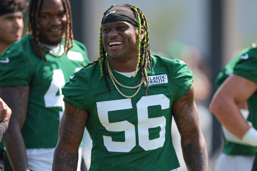 Ein lächelnder Jets-Footballspieler, Quincy Williams, der ein grünes Trikot mit der Nummer 56 und goldenen Ketten trägt, steht während des Trainings im Freien; andere Mannschaftskameraden in passenden grünen Trikots sind im Hintergrund inmitten eines defensiven Umbruchs zu sehen. Diese Beschreibung wurde mit der FootballR KI automatisch generiert.