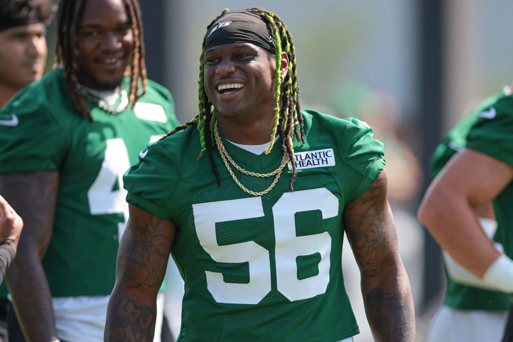 Ein lächelnder Jets-Footballspieler, Quincy Williams, der ein grünes Trikot mit der Nummer 56 und goldenen Ketten trägt, steht während des Trainings im Freien; andere Mannschaftskameraden in passenden grünen Trikots sind im Hintergrund inmitten eines defensiven Umbruchs zu sehen. Diese Beschreibung wurde mit der FootballR KI automatisch generiert.