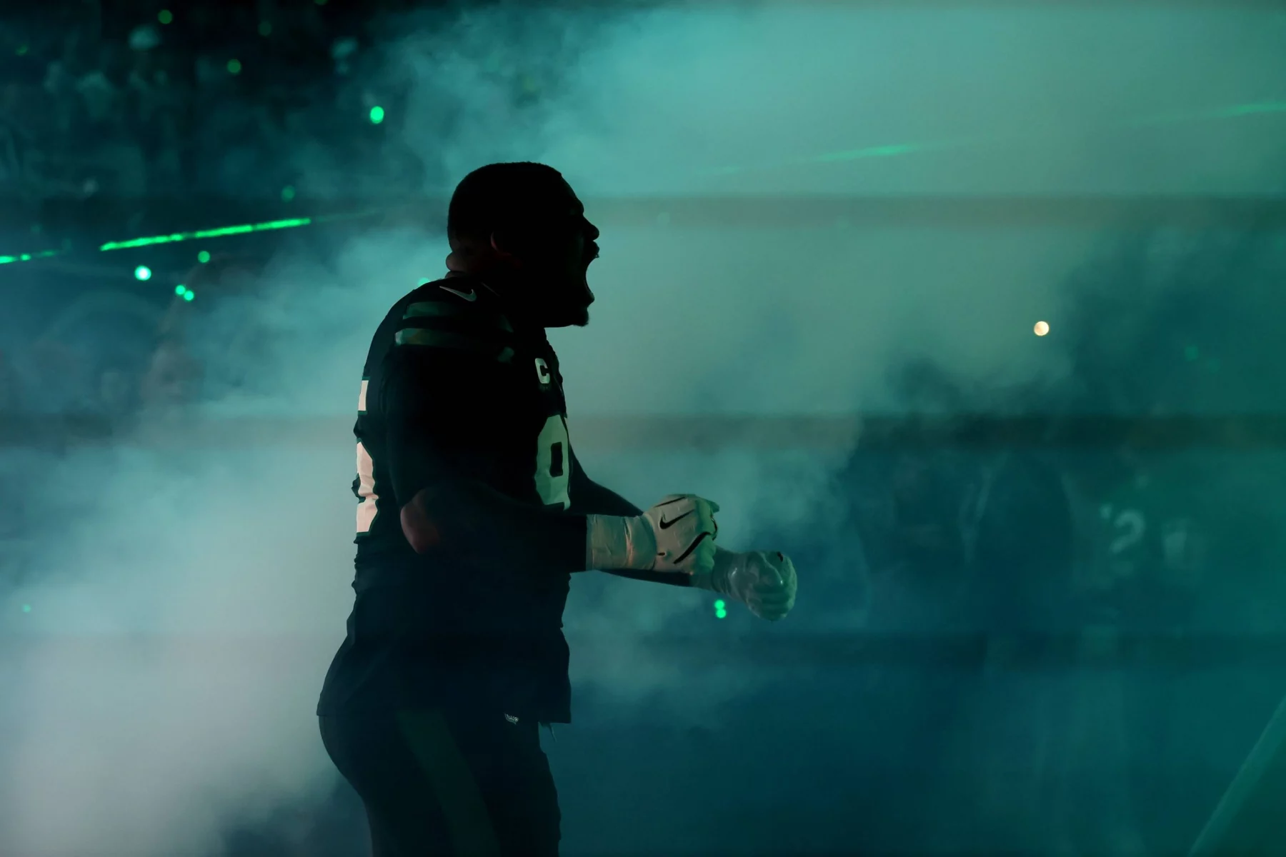 Ein Football-Spieler in voller Montur steht im Profil, schreit oder jubelt inmitten von dichtem Rauch und dramatischer grüner Beleuchtung; die intensive Atmosphäre deutet auf die Energie eines Spiels der Dallas Cowboys hin, mit undeutlichen Figuren und grünen Laserlichtern im Hintergrund. Diese Beschreibung wurde mit der FootballR KI automatisch generiert.