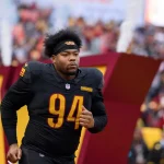 Daron Payne in einer schwarzen Uniform mit der Nummer 94, die einem Commanders Verteidiger ähnelt, läuft auf das Spielfeld. Rauch und eine große "W"-Struktur tauchen im Hintergrund auf, während die konzentrierte Menge seinen entschlossenen Auftritt verfolgt. Diese Beschreibung wurde mit der FootballR KI automatisch generiert.