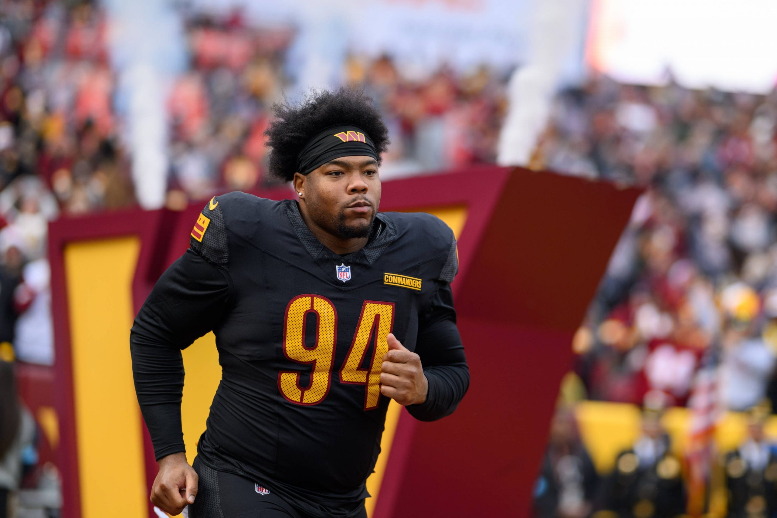 Daron Payne in einer schwarzen Uniform mit der Nummer 94, die einem Commanders Verteidiger ähnelt, läuft auf das Spielfeld. Rauch und eine große "W"-Struktur tauchen im Hintergrund auf, während die konzentrierte Menge seinen entschlossenen Auftritt verfolgt. Diese Beschreibung wurde mit der FootballR KI automatisch generiert.