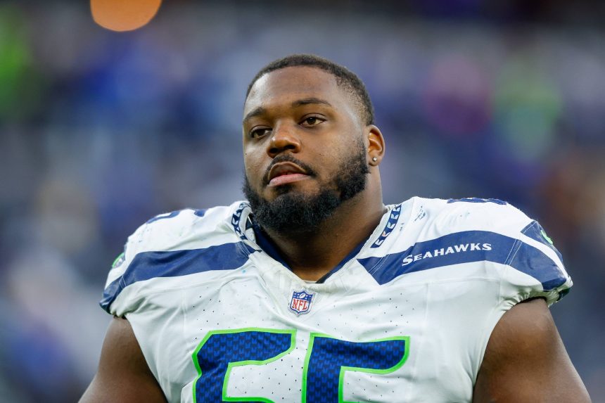 Dre’Mont Jones - Ein Football-Spieler in einem weißen Seattle Seahawks-Trikot mit der Nummer 65 steht auf dem Spielfeld. Er hat einen kurzen Bart und einen Schnurrbart, und sein neutraler Gesichtsausdruck deutet darauf hin, dass er sich darauf vorbereitet, den Pass-Rush der Ravens abzuwehren. Im Hintergrund ist eine unscharfe Stadionmenge zu sehen. Diese Beschreibung wurde mit der FootballR KI automatisch generiert.