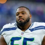 Dre’Mont Jones - Ein Football-Spieler in einem weißen Seattle Seahawks-Trikot mit der Nummer 65 steht auf dem Spielfeld. Er hat einen kurzen Bart und einen Schnurrbart, und sein neutraler Gesichtsausdruck deutet darauf hin, dass er sich darauf vorbereitet, den Pass-Rush der Ravens abzuwehren. Im Hintergrund ist eine unscharfe Stadionmenge zu sehen. Diese Beschreibung wurde mit der FootballR KI automatisch generiert.