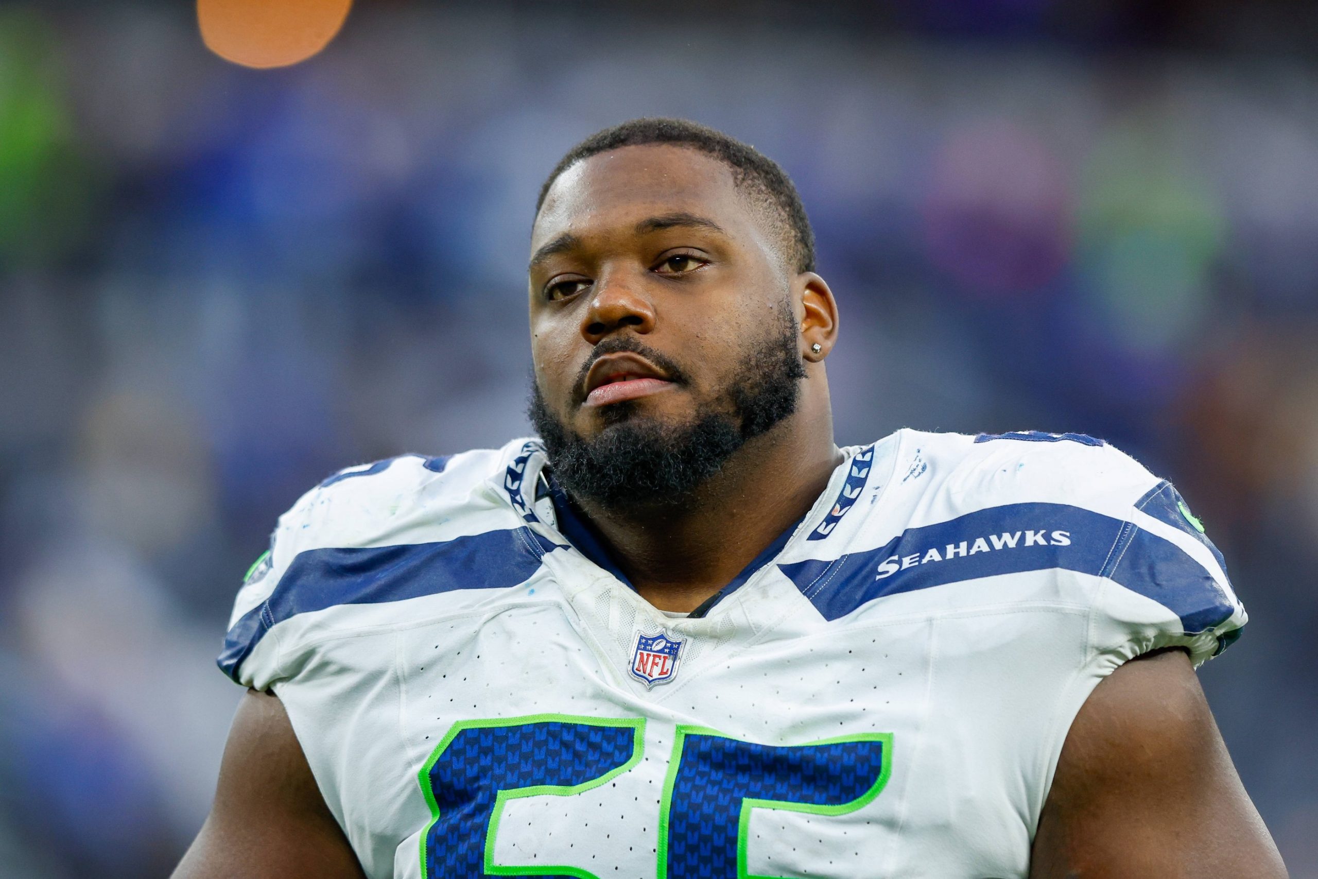 Dre’Mont Jones - Ein Football-Spieler in einem weißen Seattle Seahawks-Trikot mit der Nummer 65 steht auf dem Spielfeld. Er hat einen kurzen Bart und einen Schnurrbart, und sein neutraler Gesichtsausdruck deutet darauf hin, dass er sich darauf vorbereitet, den Pass-Rush der Ravens abzuwehren. Im Hintergrund ist eine unscharfe Stadionmenge zu sehen. Diese Beschreibung wurde mit der FootballR KI automatisch generiert.