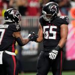 Zwei Footballspieler der Atlanta Falcons in schwarzen Trikots feiern auf dem Spielfeld in Berlin. Einer von ihnen hält einen Football in der Hand und reicht seinem Teamkollegen von der Offensive Line die Hand zum Handschlag. Im Hintergrund sind unscharfe Fans, Seitenlinienmarkierungen und Stadionlichter zu sehen. Diese Beschreibung wurde mit der FootballR KI automatisch generiert.